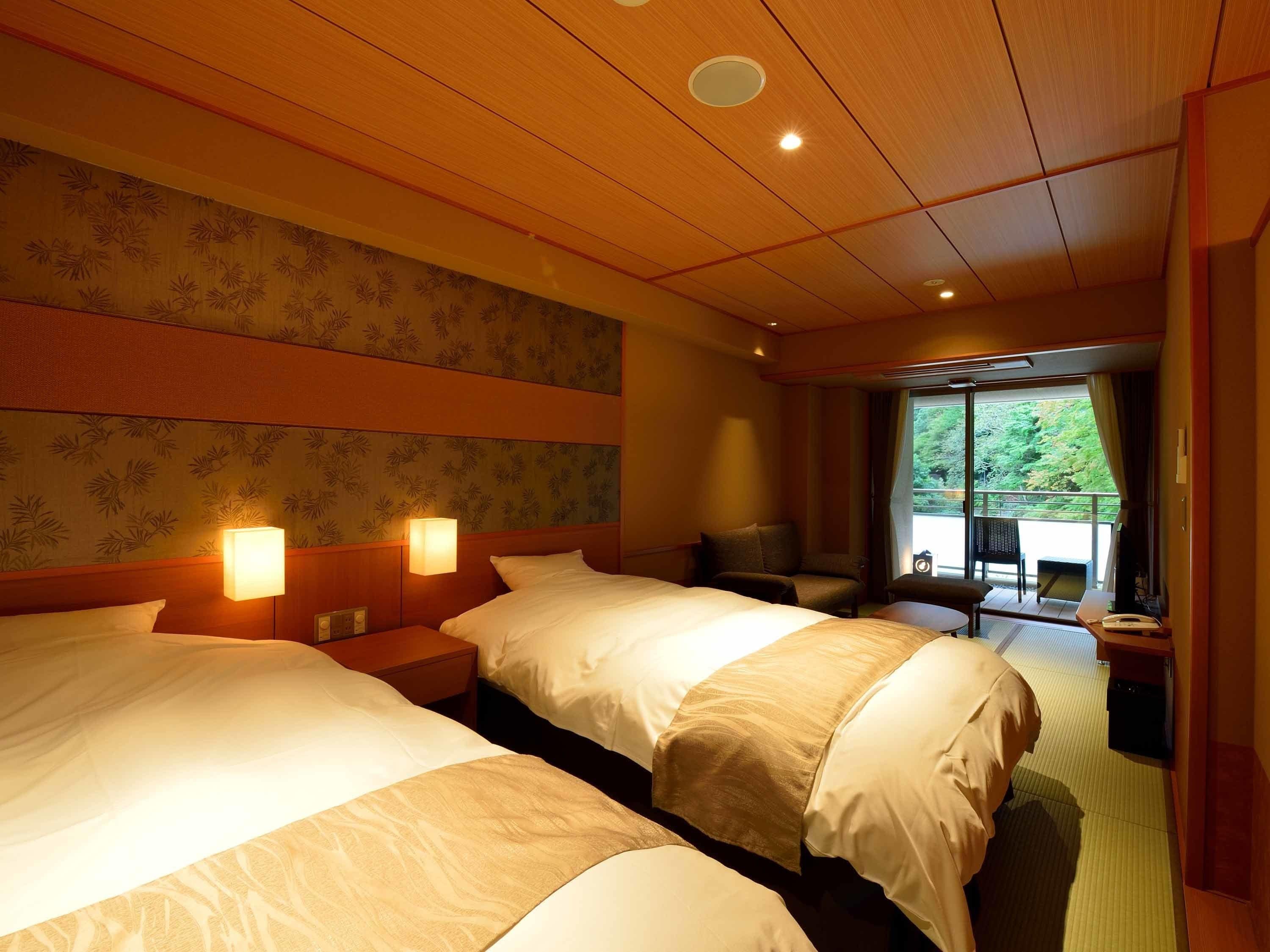 A hotel room with two beds, a desk, a chair, and a balcony with a view of greenery.