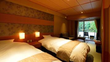 A hotel room with two beds, a desk, a chair, and a balcony with a view of greenery.
