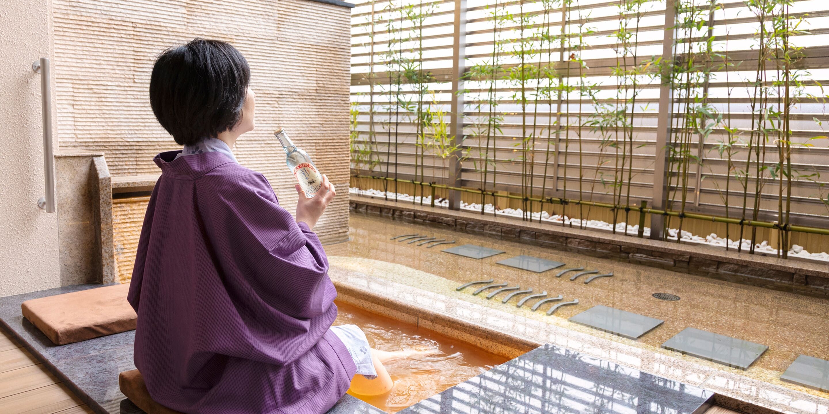 A person sitting by a hot spring with a bottle, surrounded by bamboo plants and a tiled floor.