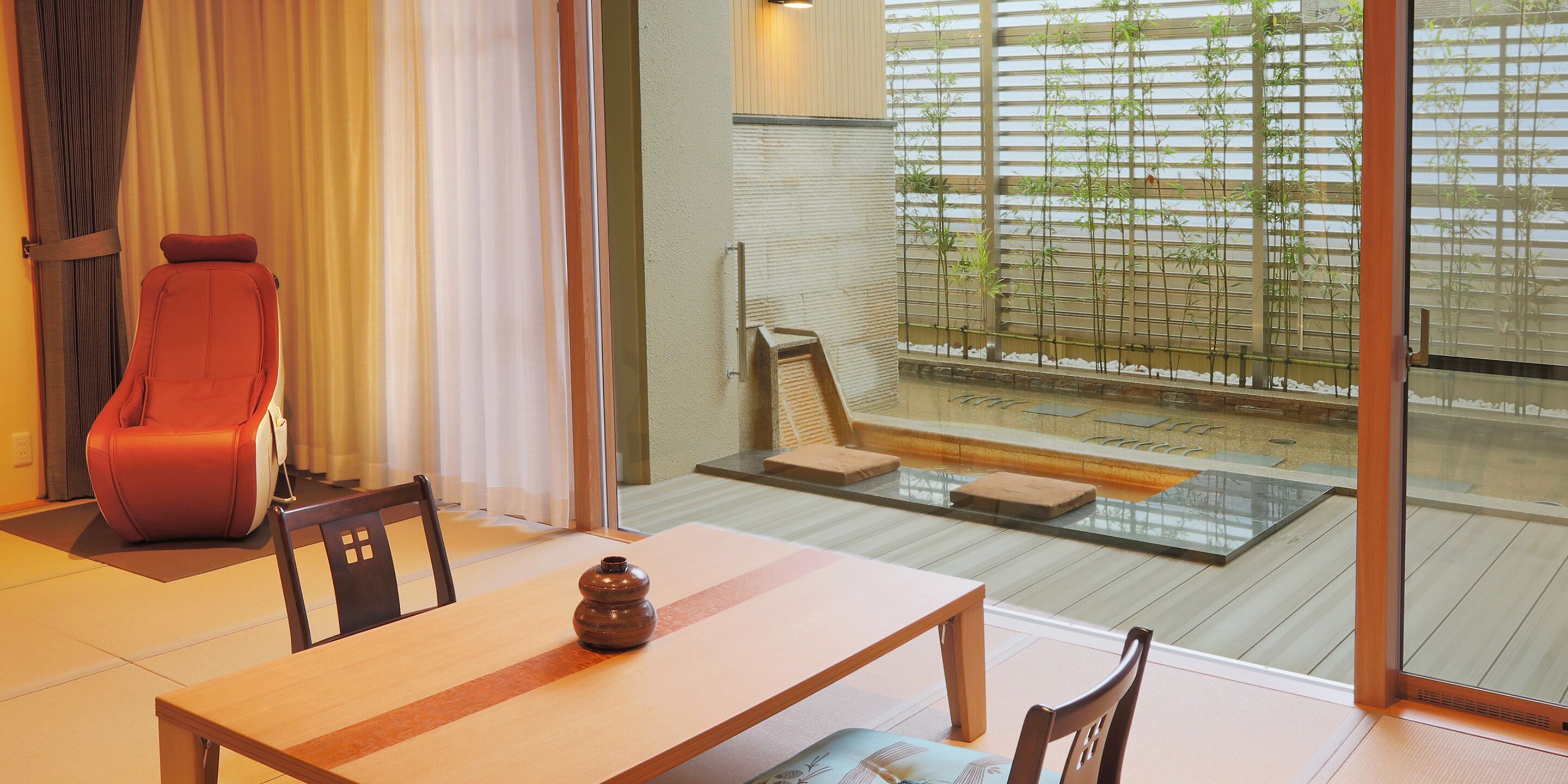 Un espace repas moderne avec une table en bois, des chaises et un fauteuil rouge. Une fenêtre offre une vue sur des plantes de bambou et une terrasse en bois à l’extérieur.