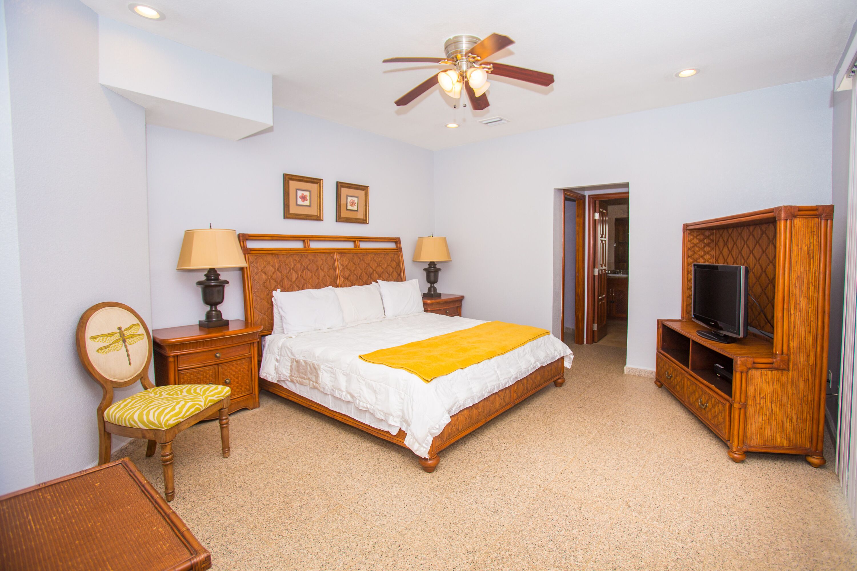 A hotel room with a bed, two bedside tables with lamps, a wooden chair, a television on a wooden stand, and a ceiling fan.