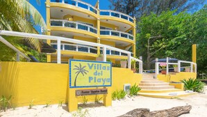 Ein gelbes Mehrfamilienhaus mit Balkonen, einer Beschilderung mit der Aufschrift "Villas del Playa" und einer Website-Adresse für Roatan Vacation Rentals.