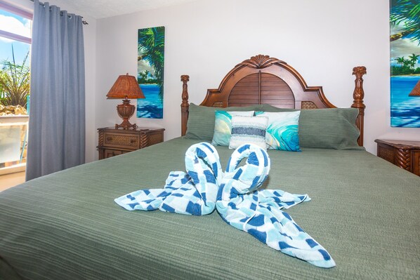 A neatly made bed with a green bedspread and blue and white patterned pillows, a wooden headboard, and two nightstands with lamps and paintings. - Villa Del Playa #1 (Roatan)