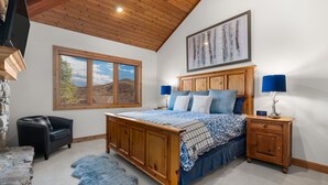 Una camera da letto con un letto di legno, un camino in pietra, una finestra con persiane, un tappeto blu e un comodino in legno con una lampada.