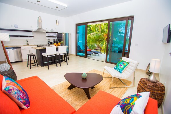 Un espacio moderno tipo sala con cocina, comedor y un amplio ventanal corredera que da a un jardín tropical.