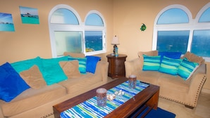 A living room with two beige sofas, a wooden coffee table, and a view of the ocean through large windows. - Stunning Coral View Home with Oceanfront Pool by RedAwning (Roatan)