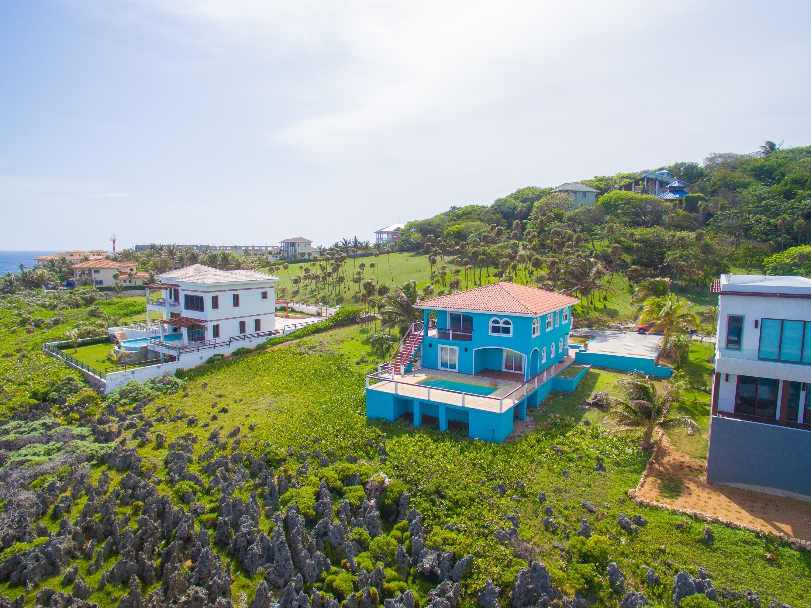 Una propiedad costera con varias construcciones, incluyendo una casa azul destacada con techo rojo, rodeada de vegetación exuberante y una vista despejada del mar.