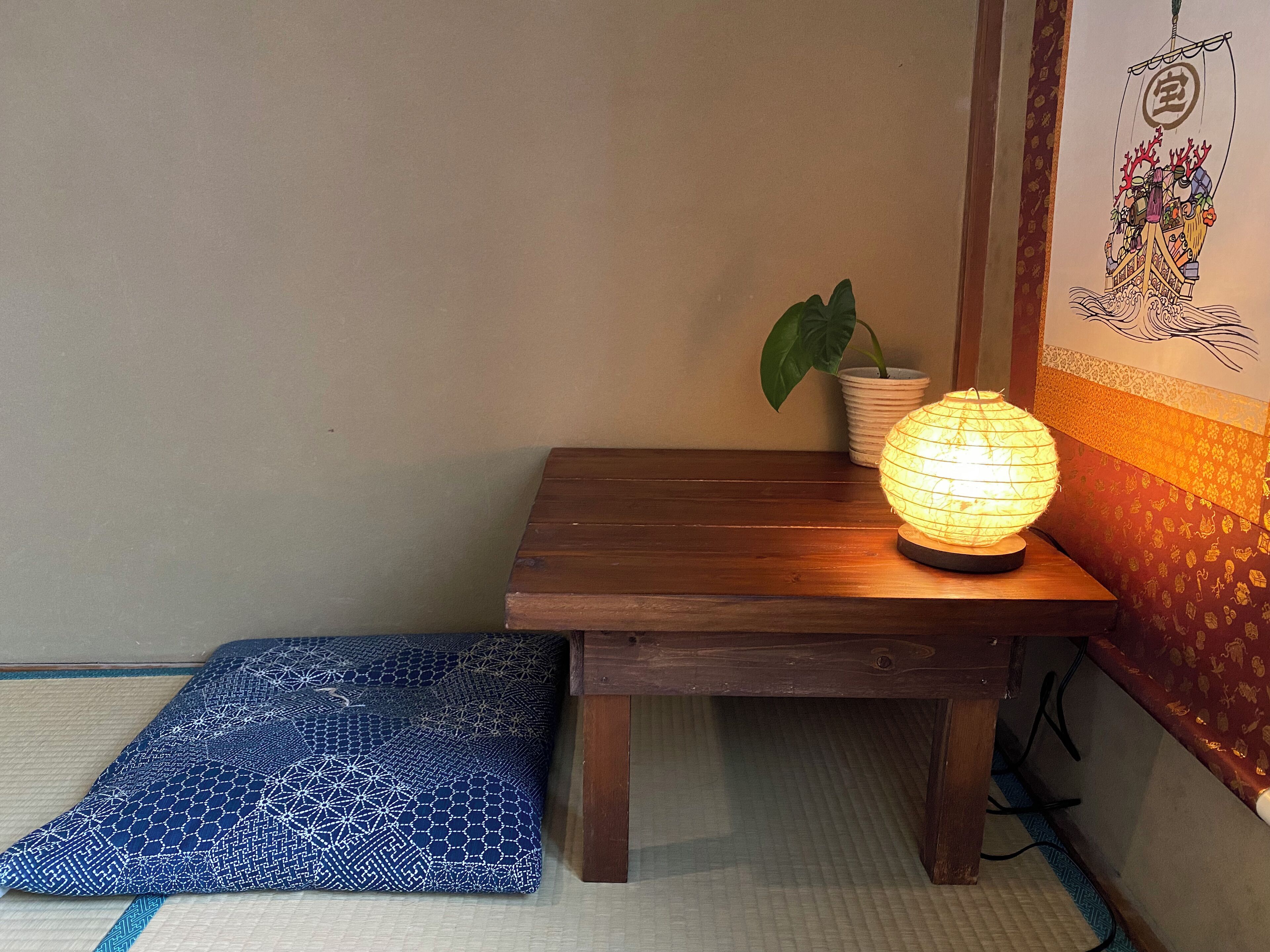 Une petite table en bois, sur laquelle est posée une lanterne en papier allumée, un pot de fleurs et un coussin bleu à motifs, le tout sur un sol recouvert de tatamis.