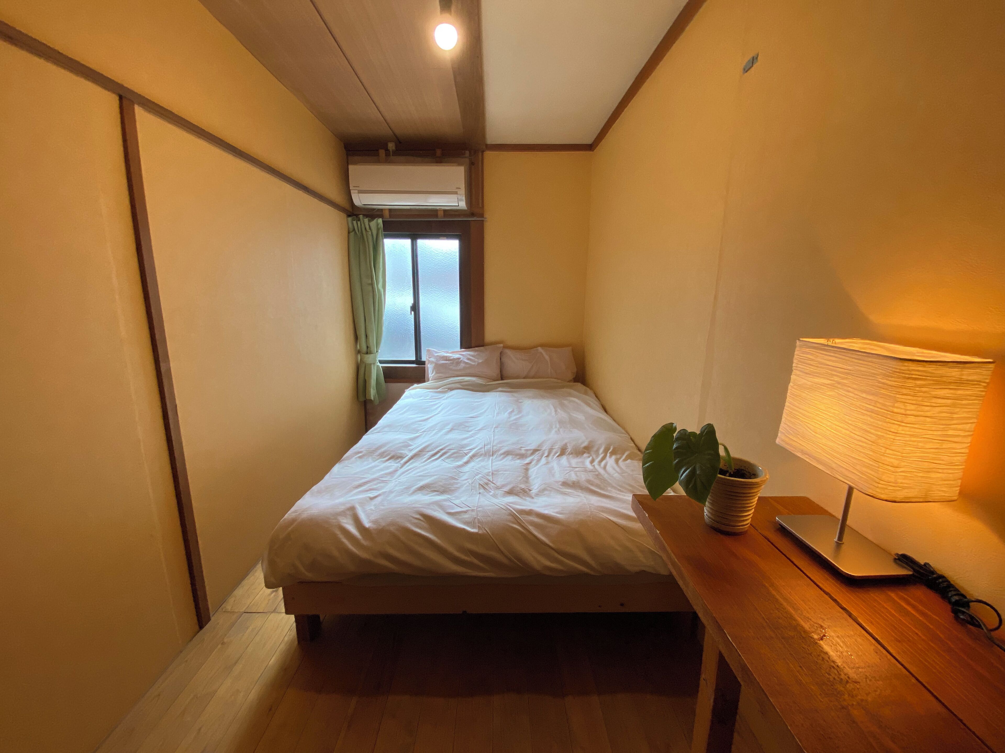A bedroom with a bed, a bedside lamp, a potted plant, and a window with curtains.