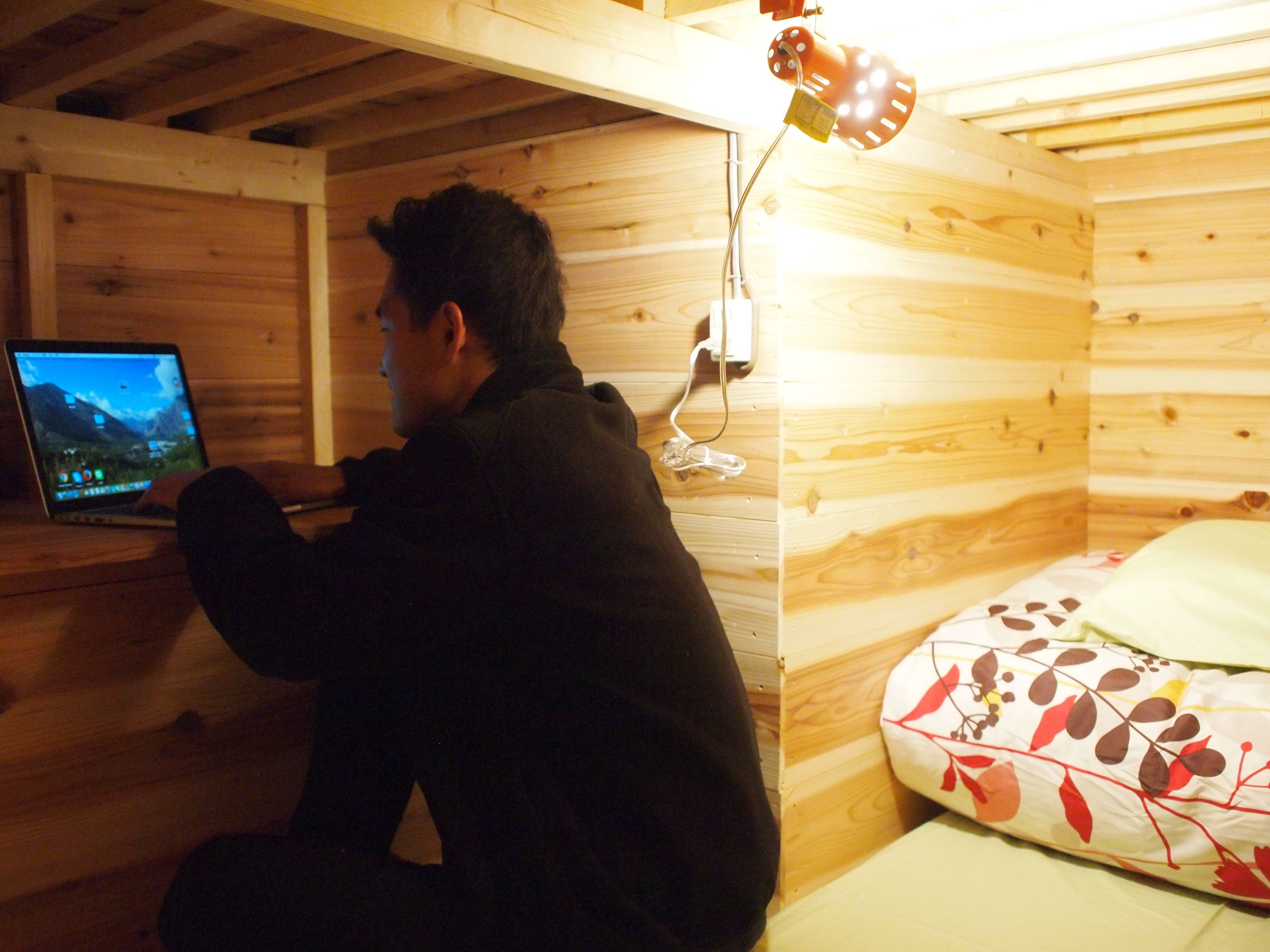 A person sitting at a wooden desk with a laptop, in a room with wooden walls and a bed with a floral pillow.