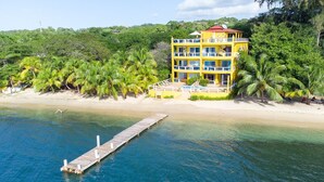 Un immeuble jaune à plusieurs étages, doté de balcons, situé sur une plage bordée de palmiers et d’une jetée qui s’avance dans l’eau.