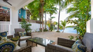 Een overdekt terras met rieten meubels en gekleurde kussens, met uitzicht op een zanderig strand en palmbomen.