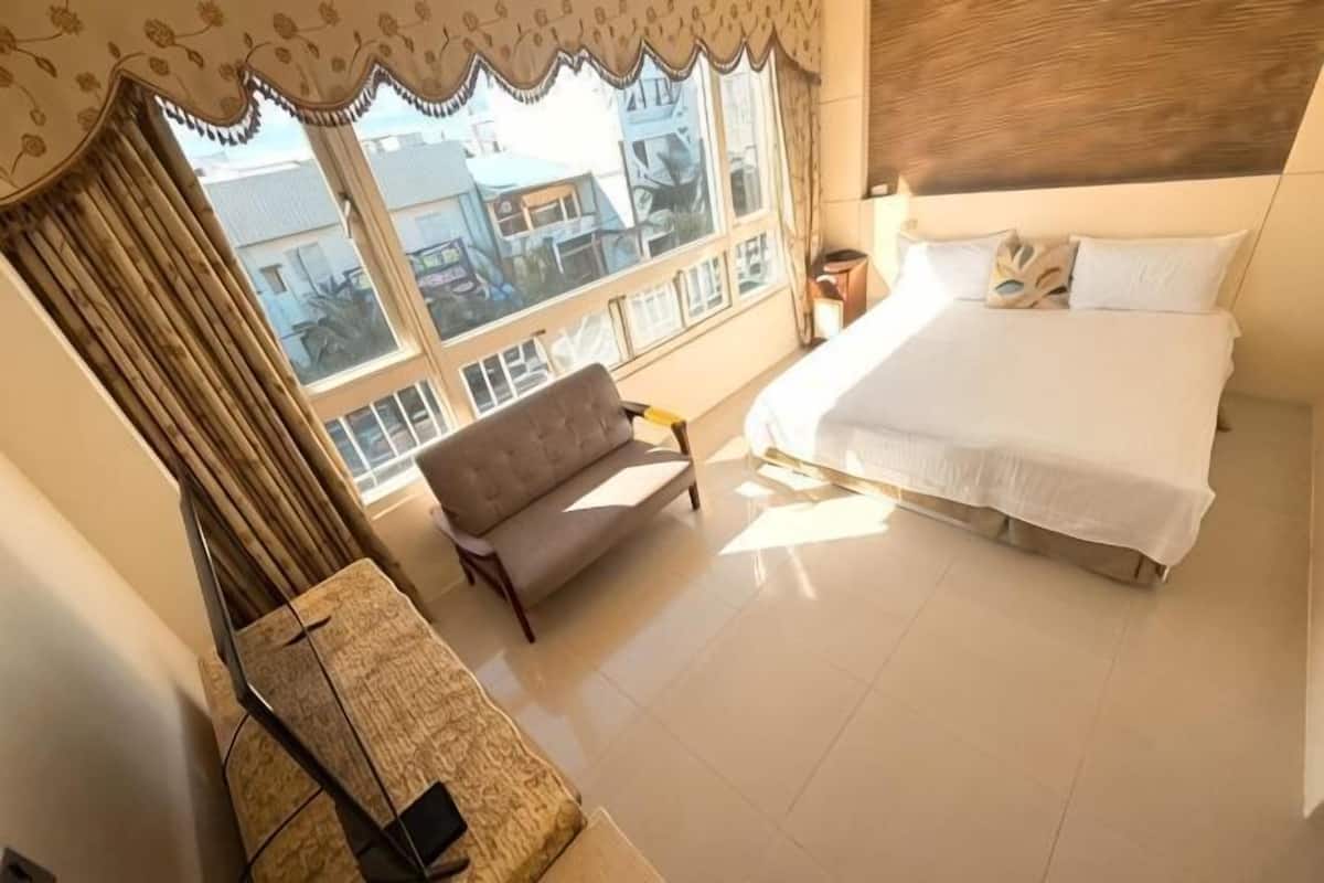 A bedroom with a bed, a sofa, a window with curtains, and a view of buildings outside.