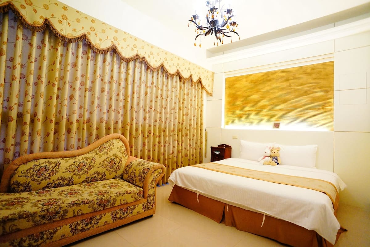 A bedroom with a sofa, a bed, a chandelier, and a window with blinds.