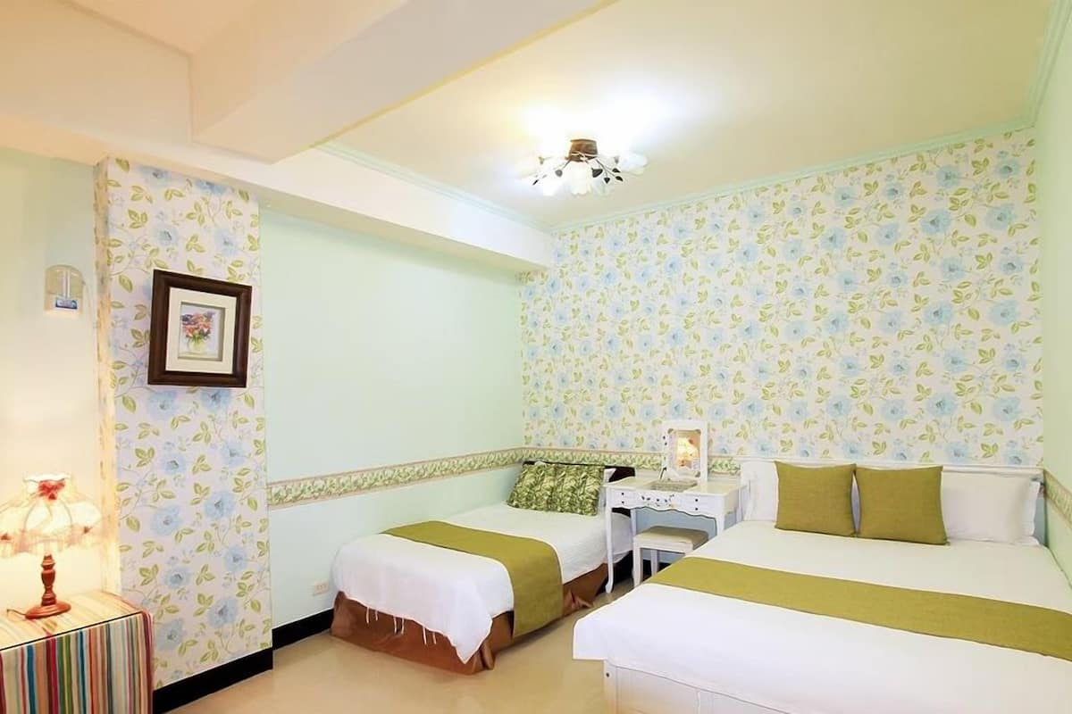 A hotel room with two beds, a bedside table with a lamp, and floral wallpaper.