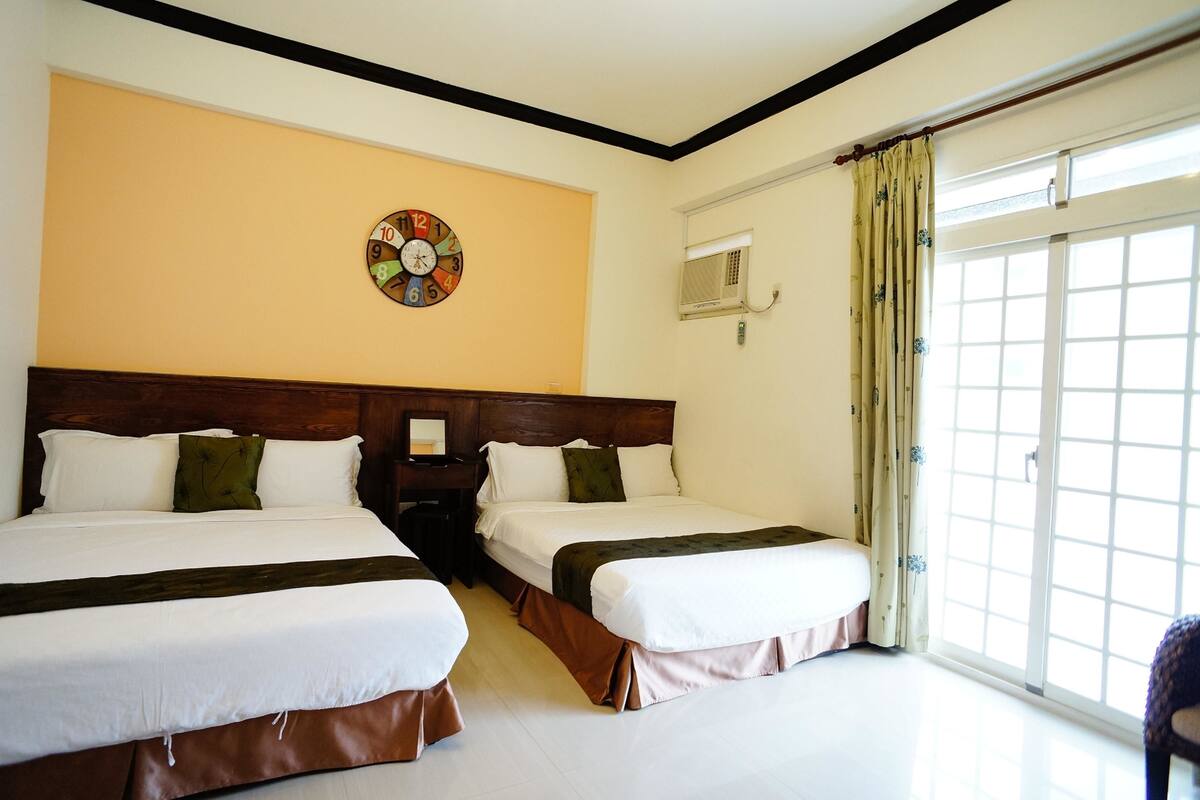 A hotel room with two beds, a wooden headboard, a wall clock, an air conditioning unit, and a large window with curtains.