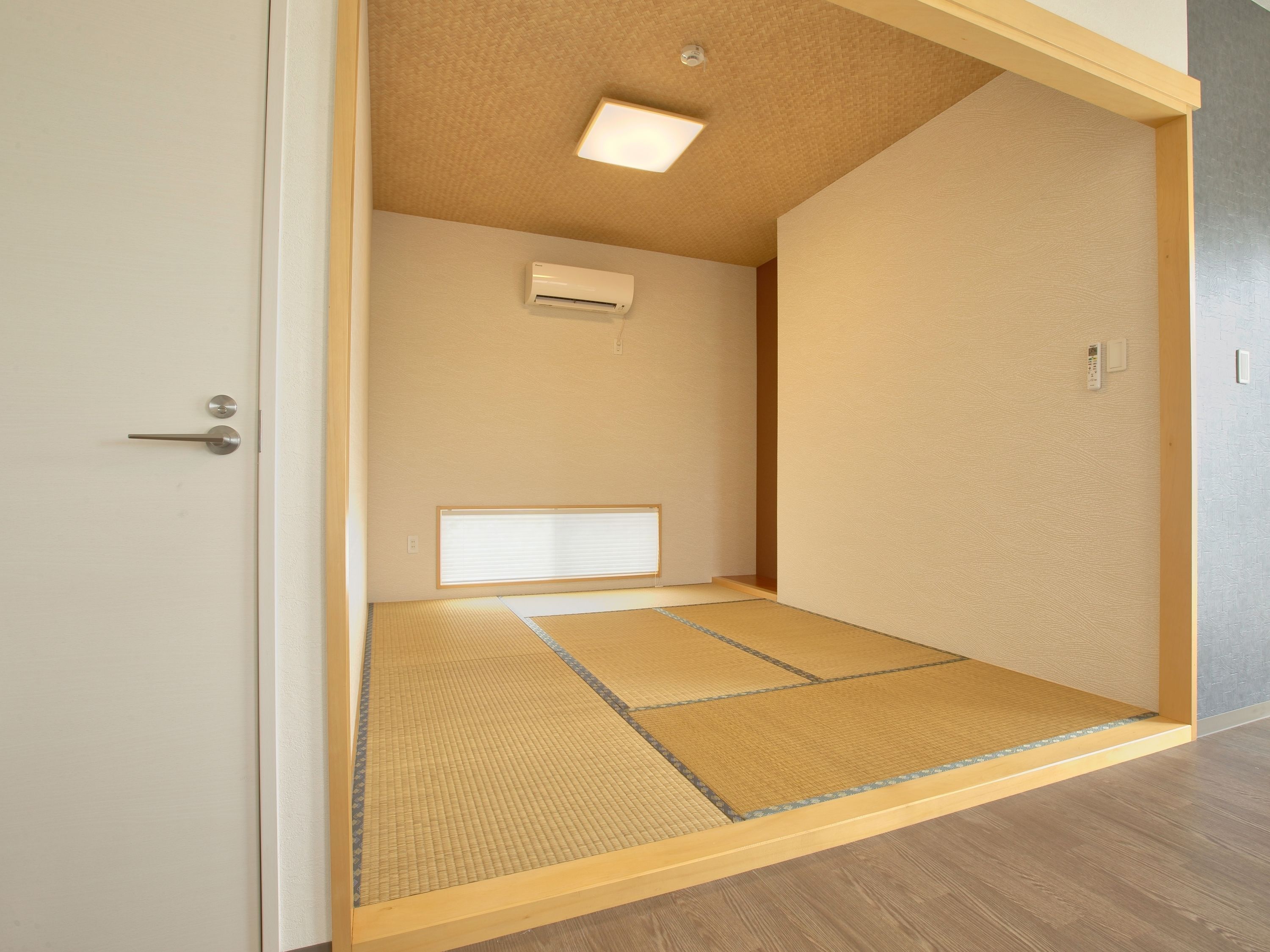 Une pièce de style japonais traditionnel, avec un sol recouvert de tatamis, une porte coulissante, un luminaire au plafond et un climatiseur fixé au mur.