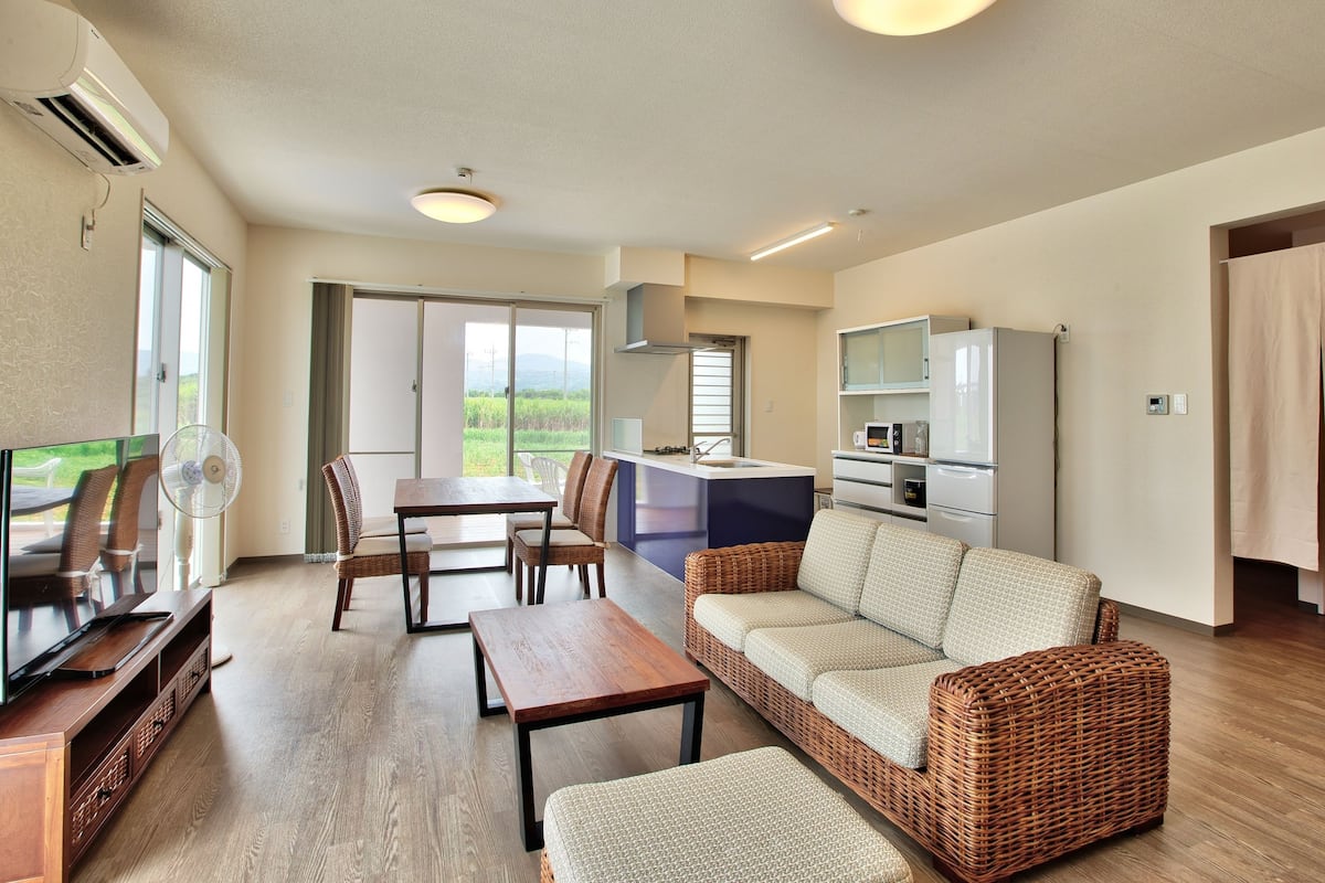 A modern living room with a sofa, a dining table, and a TV. There is a kitchen area with a refrigerator and microwave.