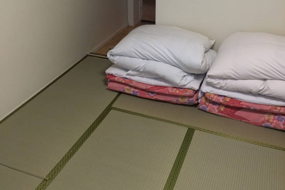 Een kamer met tatami vloermatten, een bed met witte en bloemenbedekte lakens en een houten kapstok aan de muur.
