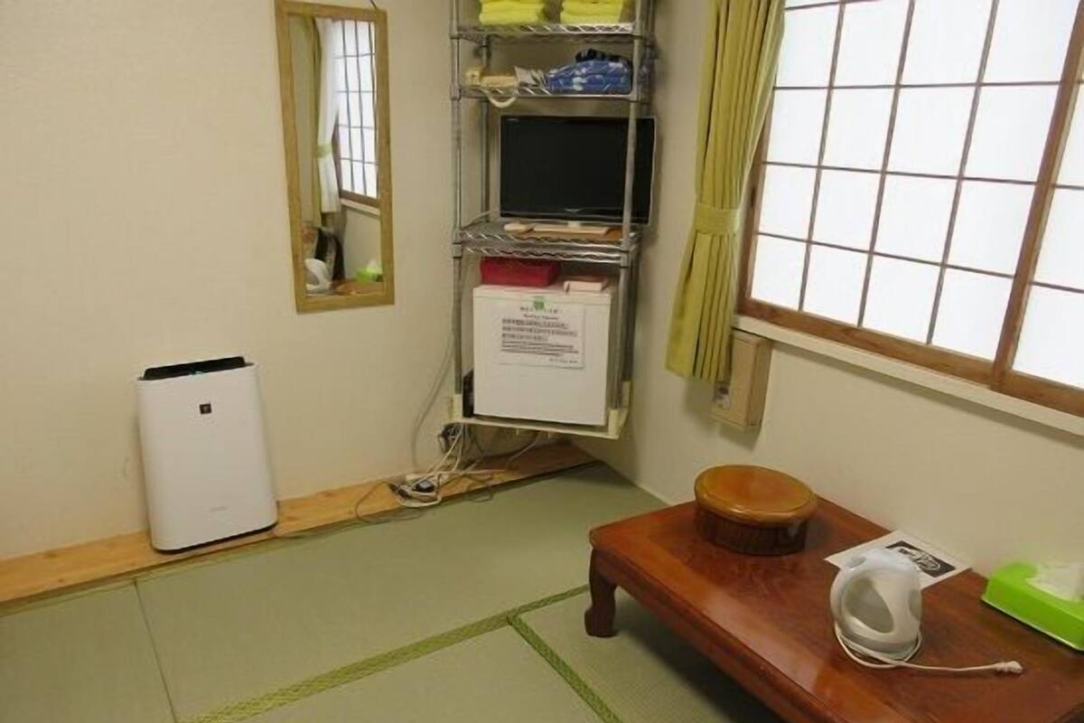 Een kamer met tatami vloerbedekking, een houten bank, een klein tafeltje met een waterkoker, een plank met opgevouwen spullen en een witte verwarming.
