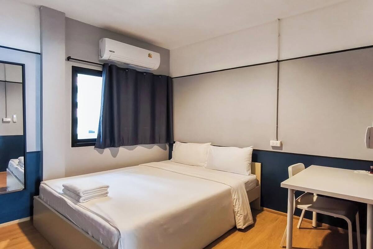 Habitación de hotel con cama, escritorio, silla, equipo de aire acondicionado y ventana con cortinas.
