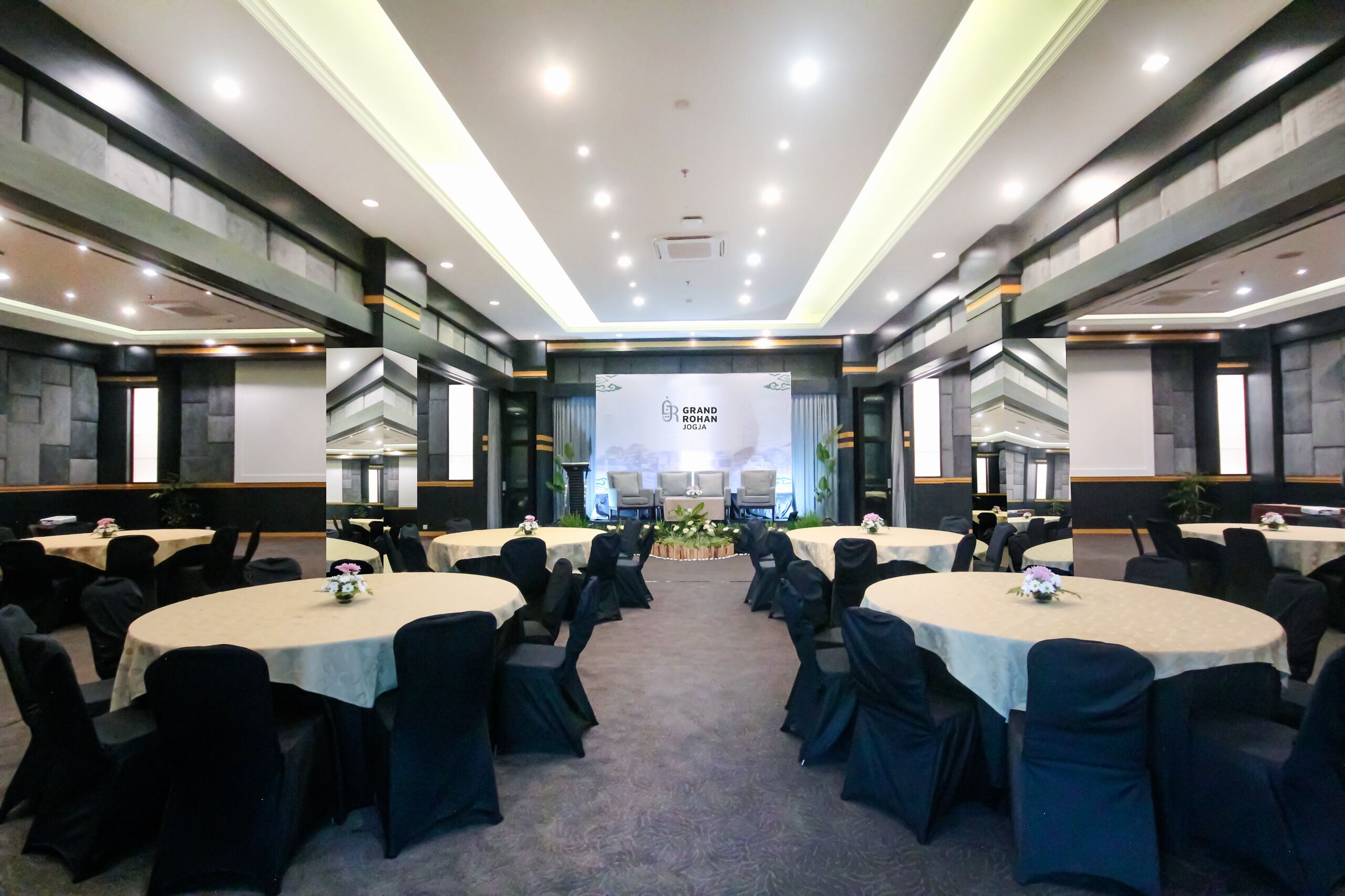 A conference room with multiple round tables, each with a white tablecloth and black chairs. The room has a modern design with a high ceiling and recessed lighting.