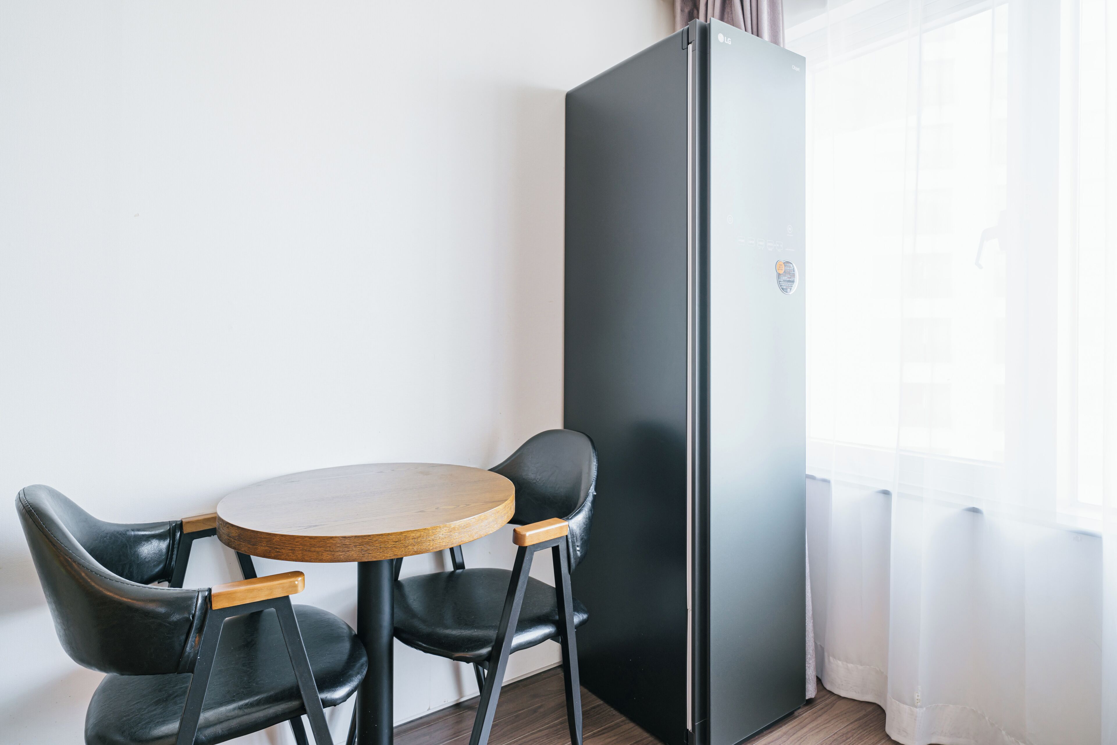 A small room with a round wooden table and two black chairs, a black refrigerator, and a window with sheer curtains.