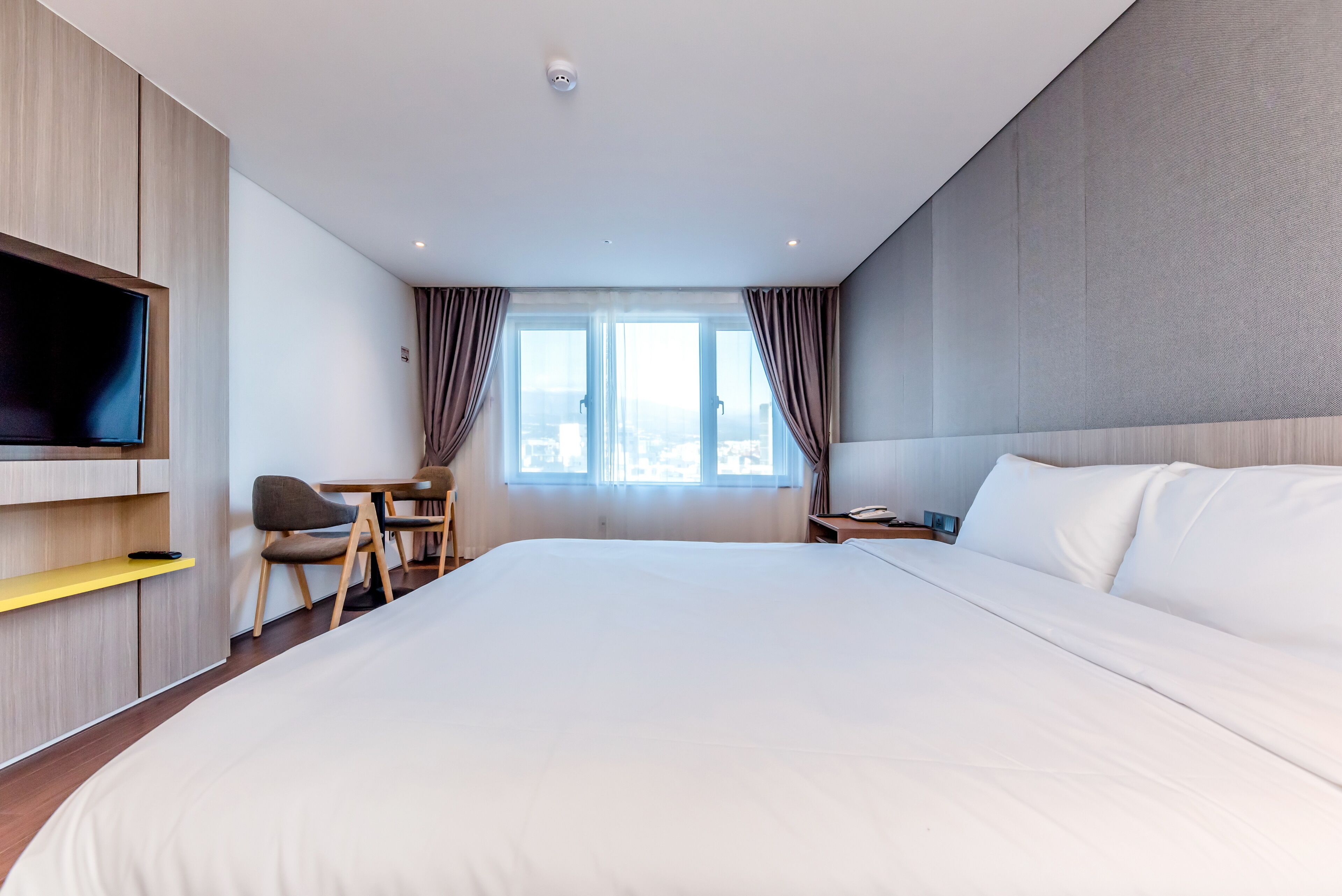 A modern hotel room with a large bed, a flat-screen TV mounted on the wall, a desk with a chair, and a window with curtains.