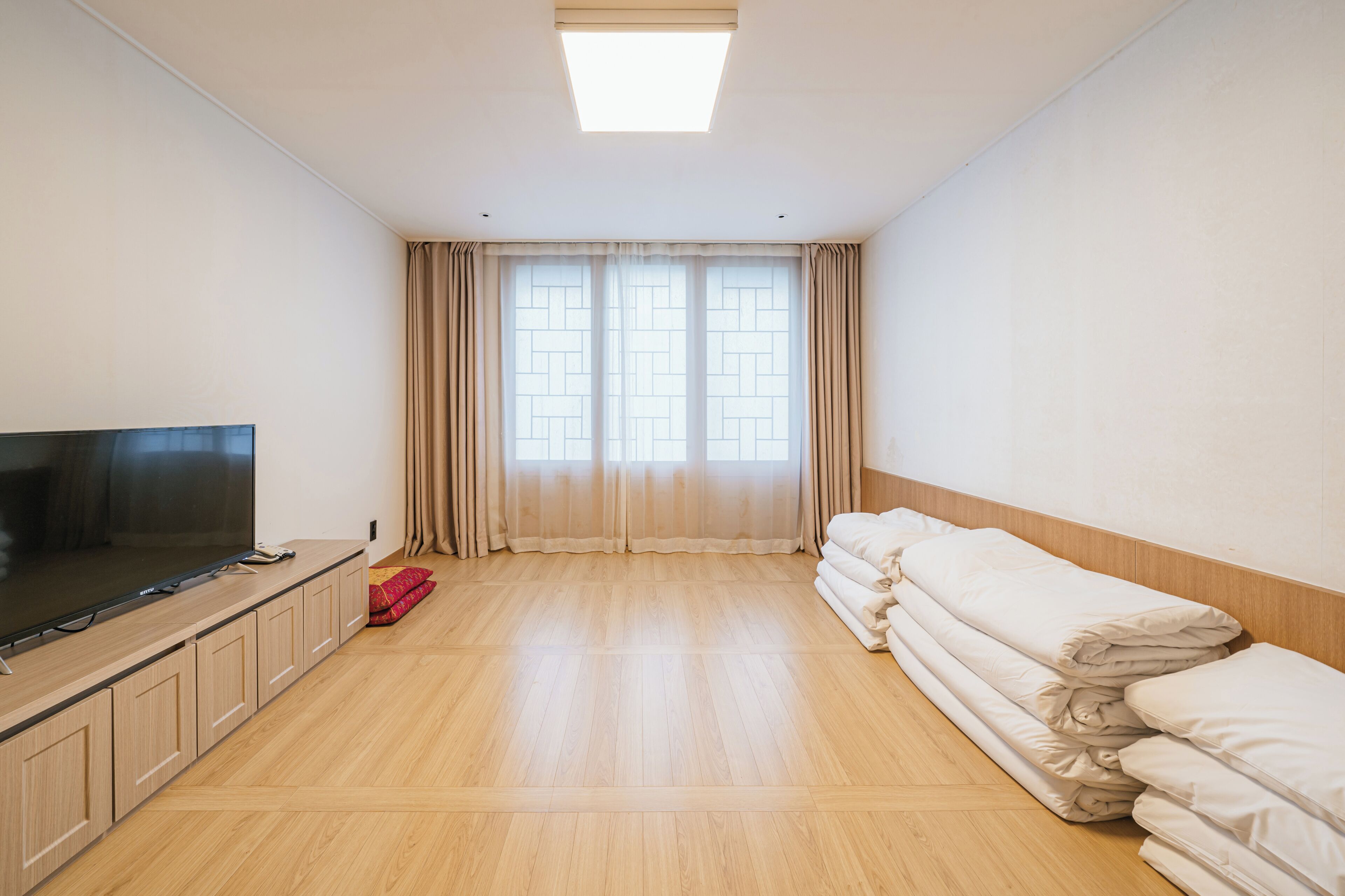 A hotel room with a flat-screen TV, a wooden floor, a large window with curtains, and neatly stacked bedding.