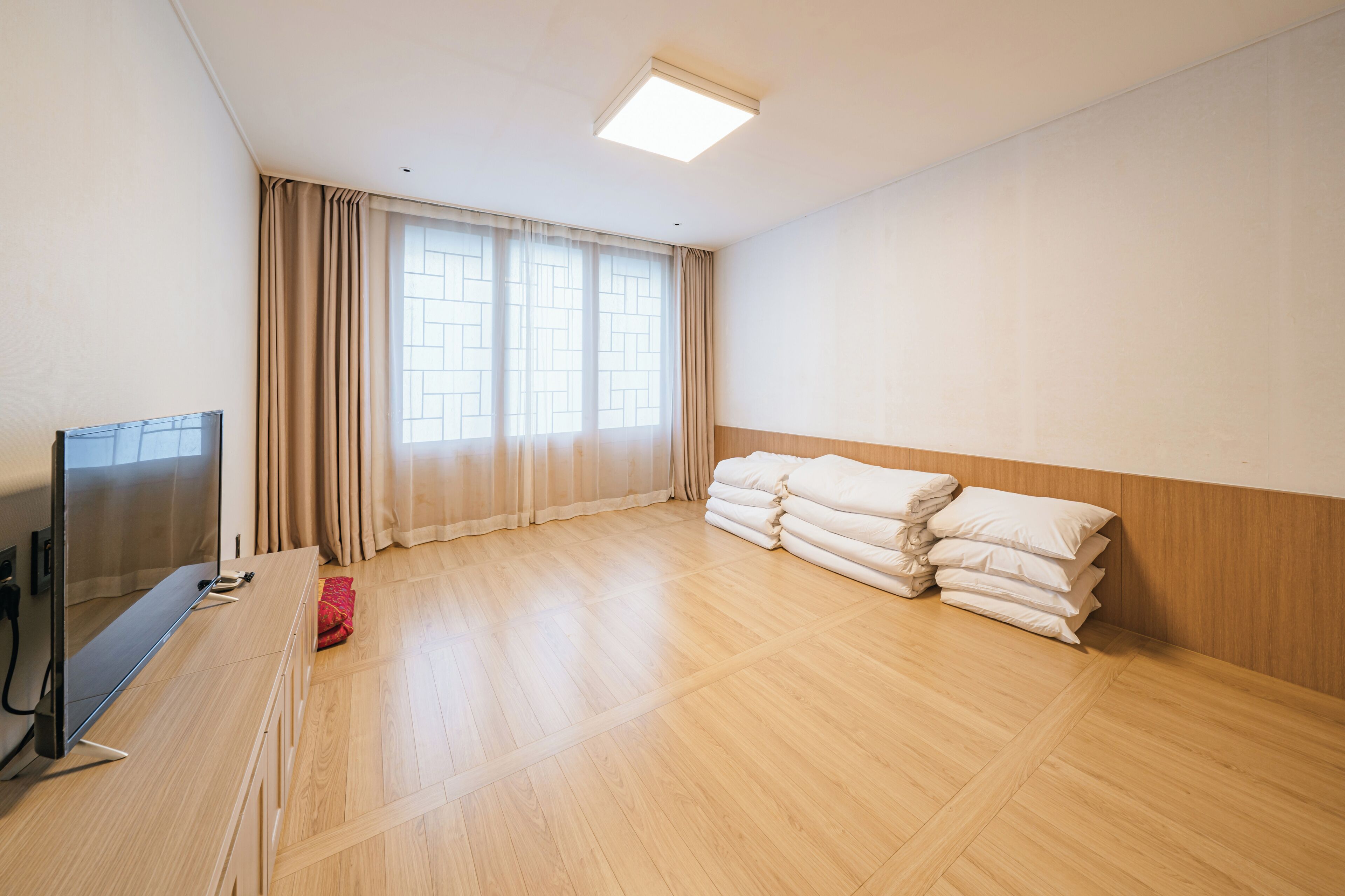A room with a wooden floor, a TV on a stand, a window with curtains, and a stack of pillows.