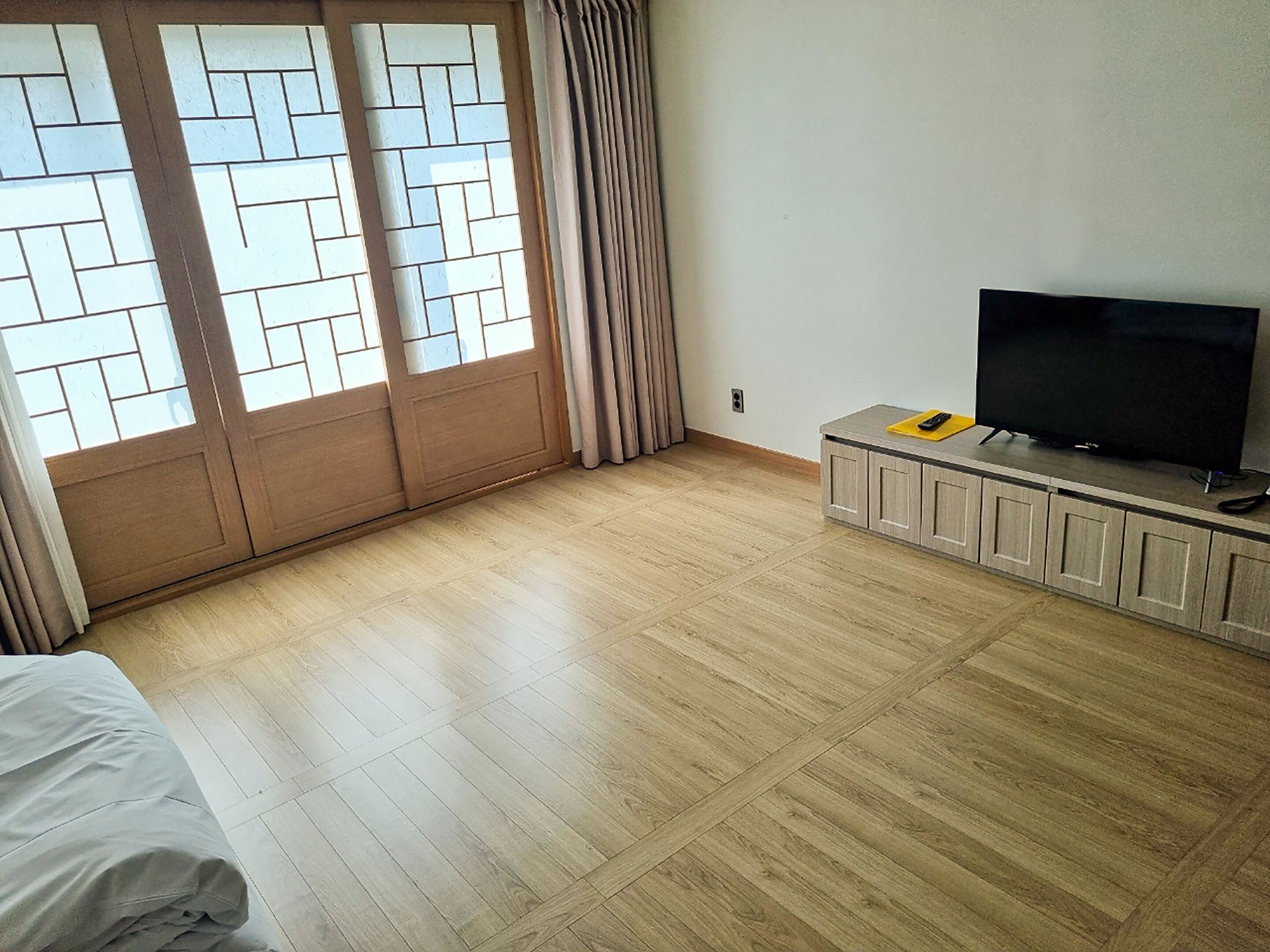 A hotel room with a large window, a flat-screen TV on a wooden cabinet, and a bed with a white cover.