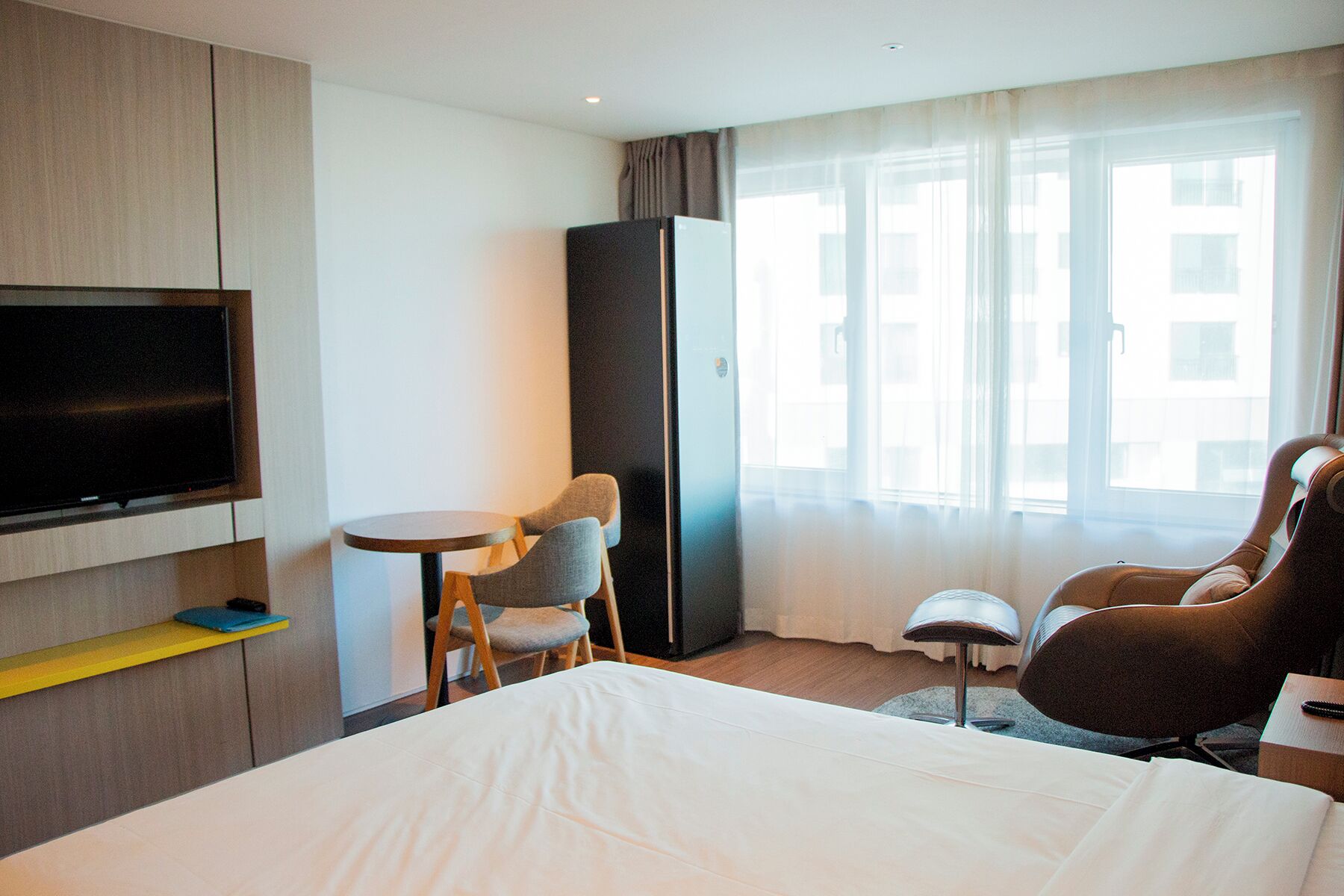 Una habitación de hotel con cama, televisión montada en la pared, una mesita redonda pequeña, dos sillas, un sofá y un ventanal con cortinas.