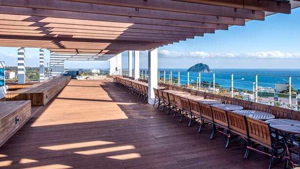 A covered outdoor seating area with wooden tables and chairs, overlooking a coastal view with buildings and a mountain in the distance. - The First70 Hotel (Seogwipo)