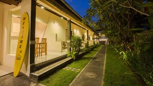 A hotel with a well-lit entrance, a surfboard leaning against the wall, and a pathway lined with greenery.