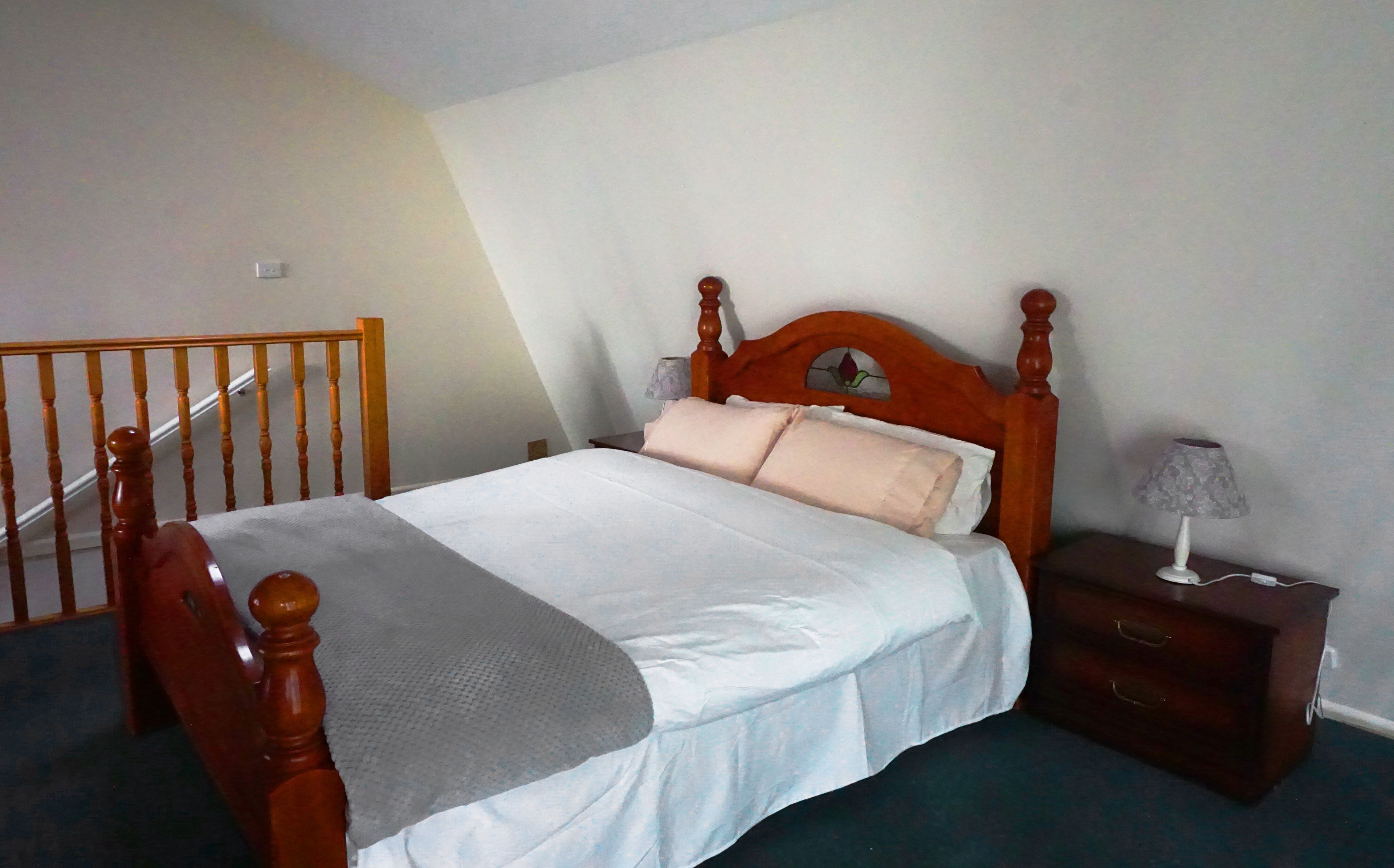 A bedroom with a wooden bed, a nightstand, a lamp, and a staircase.