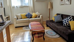 A living room with a sofa, a coffee table, and a lamp. There is a window with curtains and an air conditioning unit on the wall. - Moon House (Diamond Creek)