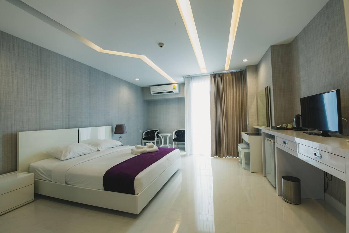 A modern hotel room with a large bed, a desk with a TV, a small seating area, and a bathroom.
