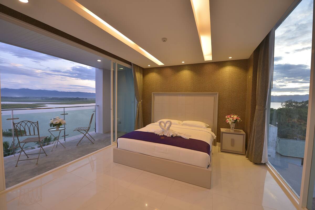 A modern bedroom with a large bed, a balcony with chairs, and a view of a body of water.