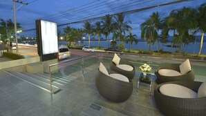 An outdoor seating area with wicker furniture and a large digital screen, overlooking a body of water and palm trees. - Phuglong Hotel (Phayao)