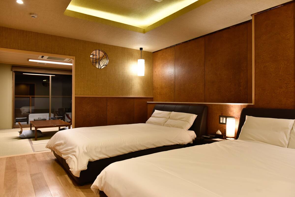 A hotel room with two beds, a wooden headboard, a bedside table with a lamp, and a sliding door leading to a sitting area.