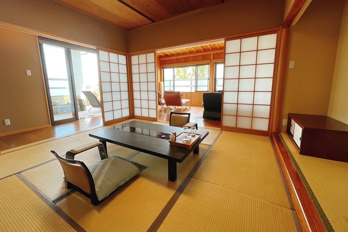 A tatami mat room with a low table and chairs, sliding doors, and a view of the outdoors.