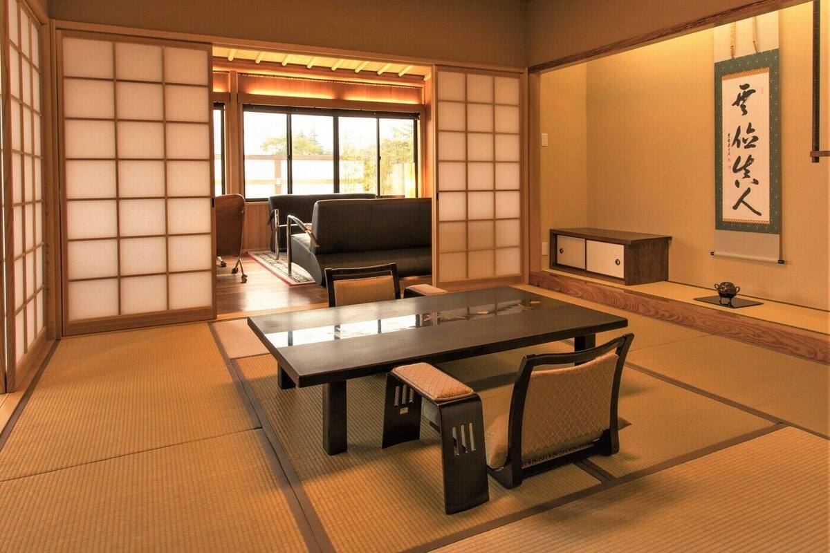 A tatami mat room with a low table and chairs, sliding doors, and a view through a window.
