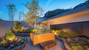 Un área de spa moderna al aire libre con un jacuzzi de madera, rodeado por un jardín de rocas y diversas plantas.