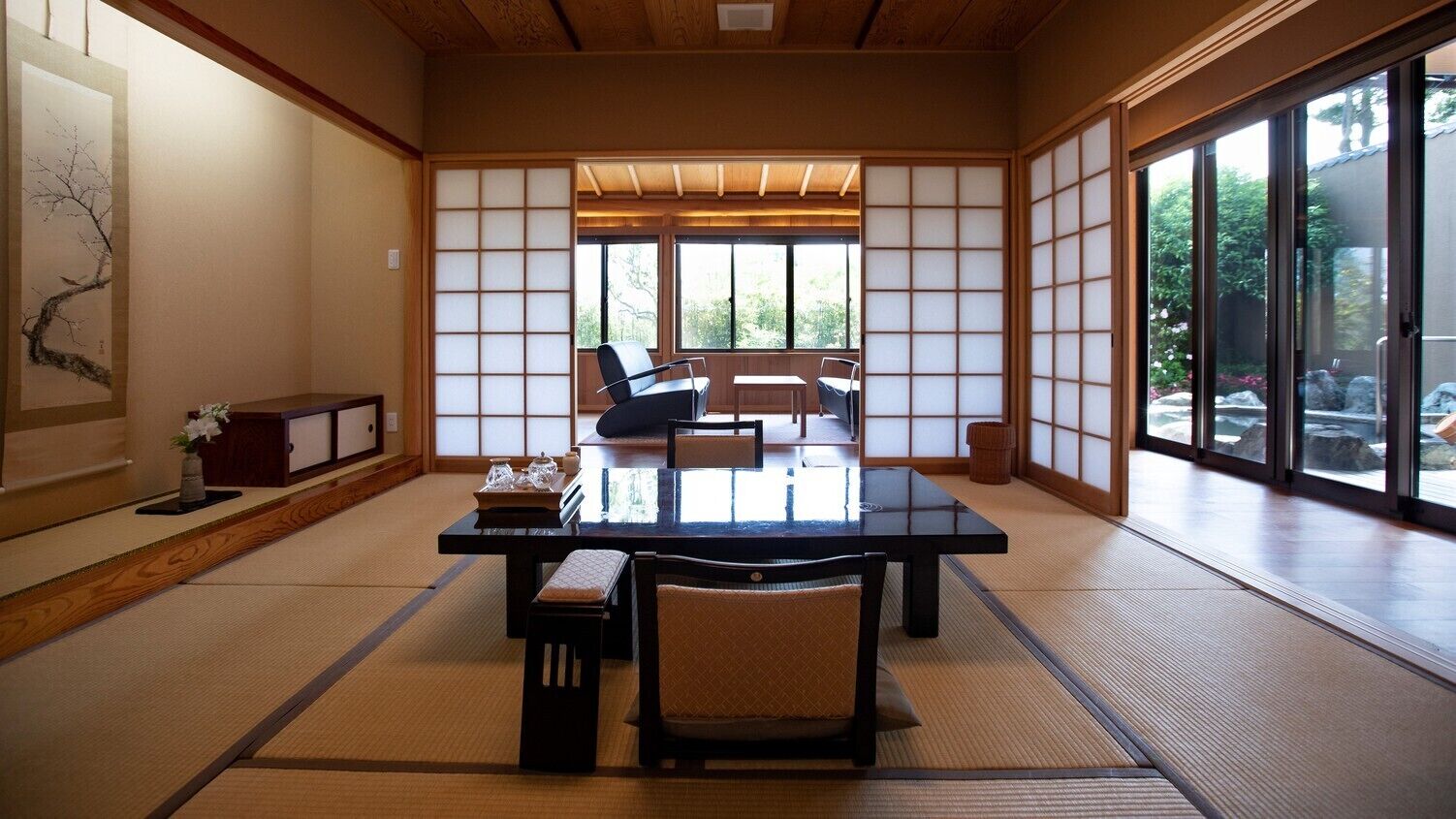 A traditional Japanese room with tatami flooring, a low table with chairs, sliding doors, and a view through large windows.