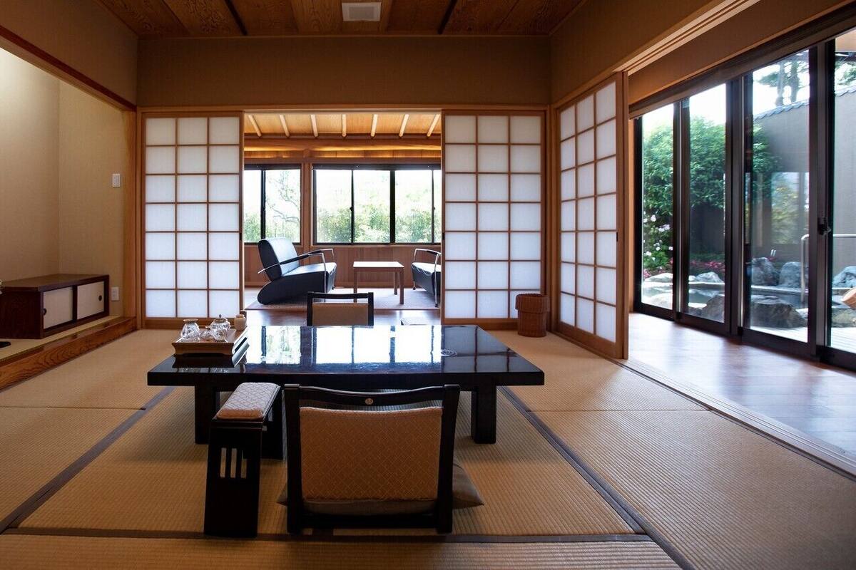 A traditional Japanese room with tatami flooring, a low table with chairs, sliding doors, and a view through large windows.
