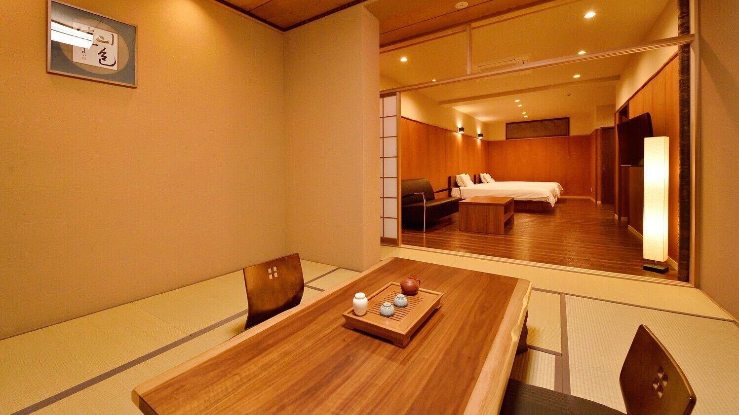 A hotel room with a wooden dining table, a traditional Japanese tea set, a bed with white linens, and a sliding door leading to another room.