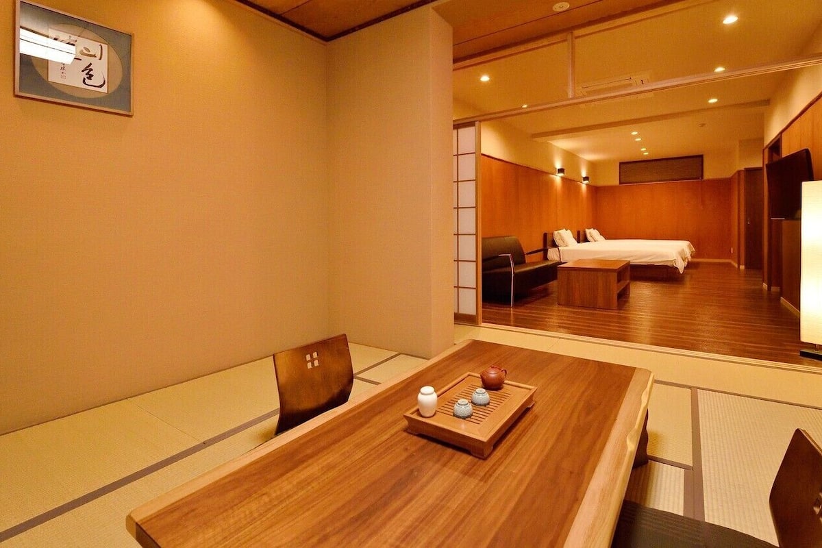 A hotel room with a wooden dining table, a traditional Japanese tea set, a bed with white linens, and a sliding door leading to another room.