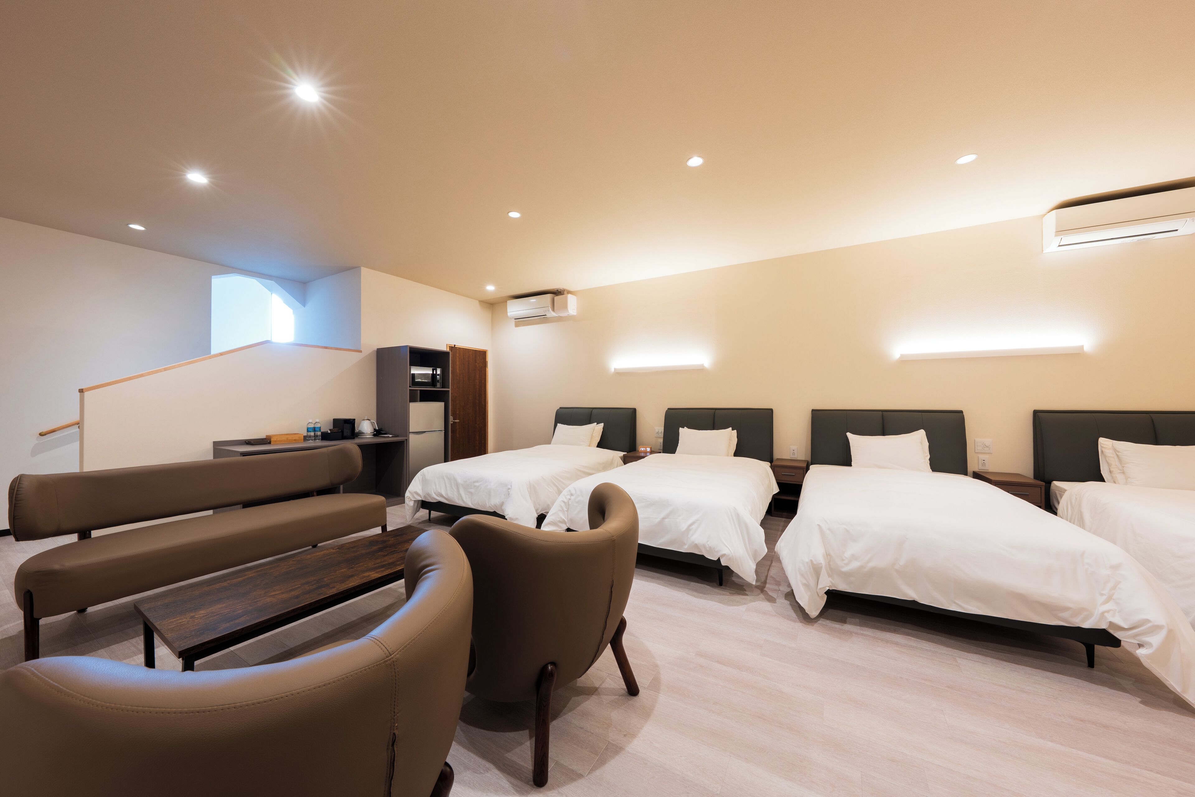 A hotel room with two beds, a bench, a chair, a small table, and a kitchenette.