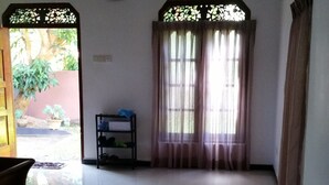 A room with a wooden bench, a small shelf with shoes, a clock on the wall, and a window with curtains. - Srilusa Home Stay Hikkaduwa (Hikkaduwa)
