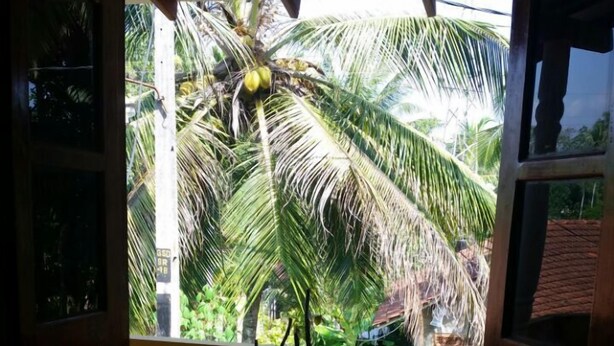 A room with a bed, a wooden table, and a view of a balcony with palm trees. - Srilusa Home Stay Hikkaduwa (Hikkaduwa)